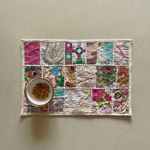 May include: A rectangular, patchwork placemat with a variety of embroidered fabric squares in pink, blue, green, and cream. A small white bowl with a gold rim holds a serving of almonds. The placemat is bordered with a white fabric.