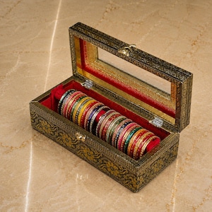 Könnte beinhalten: Eine rechteckige Schmuckschatulle mit Klappdeckel und Sichtfenster, die ein schwarzes Äußeres mit goldenen Blumenmustern und ein rotes Samtfutter aufweist. Im Inneren ist eine Reihe dünner, mehrfarbiger Metallarmreifen hintereinander aufgereiht.