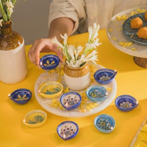 Könnte beinhalten: Eine Sammlung kleiner, handbemalter Keramik-Öllampen in verschiedenen Farben, darunter Blau und Gelb, angeordnet auf einem weißen Marmortablett. Jede Lampe hat Blumendesigns und eine kleine Flamme, mit weißen Blumen in einer Vase.