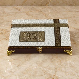 Peut inclure: Une boîte rectangulaire présentant une surface blanche avec un motif circulaire répétitif, accentuée par des bandes décoratives marron foncé. Une plaque centrale en métal doré à motif floral est présente, ainsi que des pieds d'angle dorés.