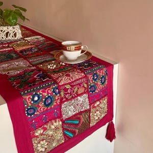 May include: A vibrant, patchwork table runner in shades of red, gold, and blue. The runner features intricate embroidery and a tasseled edge. A white teacup with gold trim sits on a matching saucer. A small potted plant is visible in the background.