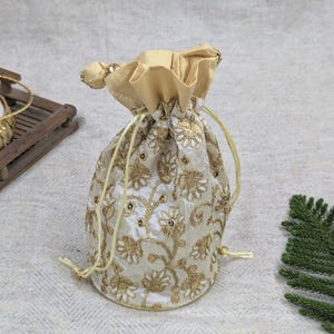 Può includere: Un sacchetto con coulisse color crema con intricati ricami floreali dorati. Il sacchetto è raccolto nella parte superiore con un tessuto dorato abbinato e una chiusura con coulisse. Un accento decorativo di perline dorate è in cima.