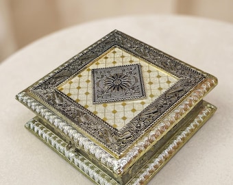 May include: A decorative square box with ornate silver and gold detailing. The lid features a central square with a floral design, surrounded by a patterned border. The box is likely used for jewelry or keepsakes.