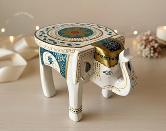 May include: A white wooden elephant stool with intricate gold and blue painted details. The round top features a floral design, and the elephant's head forms the front of the stool. The legs are painted white with gold accents.