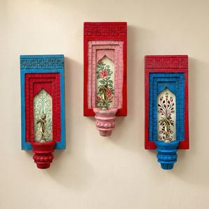 Puede incluir: Tres apliques de pared decorativos con un diseño tallado y colorido. Cada aplique presenta un marco rectangular en rojo y azul, que rodea una escena floral pintada y una pequeña estatua dorada. Los apliques están montados en una pared de color claro.