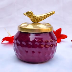 Peut inclure: Un pot décoratif avec un corps texturé, violet foncé et un couvercle doré. Une petite figurine d'oiseau dorée est posée sur le couvercle. Des pétales de fleurs rouges sont dispersés autour du pot sur une surface blanche.