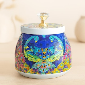 Peut inclure: Un pot décoratif en céramique avec un couvercle, orné d'un motif de paon vibrant dans les tons de bleu, vert, jaune et rose. Le couvercle a un bouton en cristal transparent et une base dorée. Le pot est blanc à la base et sur le bord.