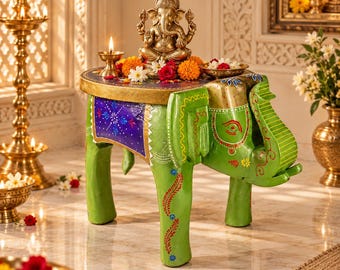 May include: A green elephant-shaped side table with a round gold top. The table features hand-painted floral patterns in red, blue, and yellow. A gold-toned Ganesha statue sits on the table, surrounded by orange and white marigold flowers.