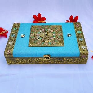 May include: Rectangular turquoise box with ornate gold-colored metal trim and a peacock design on the lid. The box has a textured surface and is adorned with floral accents and a decorative clasp.