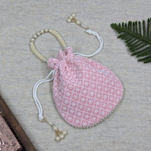 Op de afbeelding: Een roze trekkoordzakje met een wit bloemmotief. Het zakje heeft een met parels versierde handgreep en een witte trekkoordsluiting. Een decoratieve kwast met parels hangt aan het trekkoord. Het zakje wordt waarschijnlijk gebruikt voor het dragen van kleine items.