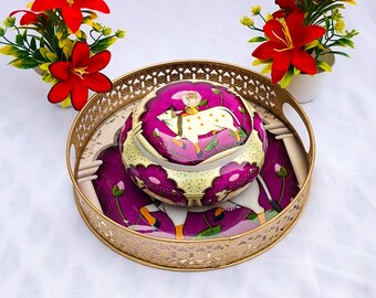 May include: A decorative gold-colored metal tray with a matching round container featuring a purple and white design. The container has a lid with a gold knob. Two small white pots with red flowers are on either side of the tray.