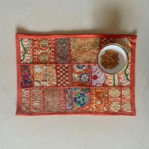 May include: A rectangular, patchwork-style placemat with an orange border. The mat features a variety of embroidered fabric squares in shades of orange, gold, and red. A small white bowl filled with almonds sits on the mat.