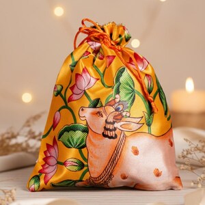 Peut inclure: Pochette jaune à cordon de serrage avec une illustration détaillée d'une vache, de fleurs de lotus et de feuilles vertes. Le sac est en tissu brillant et possède un cordon de serrage orange. L'arrière-plan est flou avec un éclairage doux.