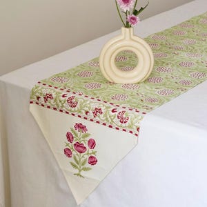 Peut inclure: Chemin de table floral vert et blanc avec un vase crème contenant des fleurs roses. Le chemin de table présente un motif répété de fleurs roses sur fond vert et une bordure décorative avec des accents rouges. Le chemin de table est sur une nappe blanche.