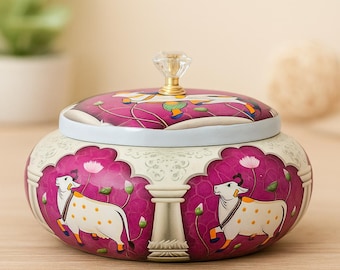 May include: A decorative, lidded ceramic jar with a vibrant design. The jar features a magenta and white floral pattern with two white cows with orange spots. The lid has a crystal knob and gold base. The jar is on a wooden surface.