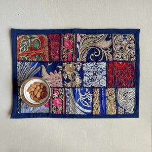 Op de afbeelding: Een rechthoekige placemat in patchwork-stijl met een marineblauwe rand. De patches zijn voorzien van ingewikkelde borduursels in rood, goud, zilver en blauw. Een kleine kom met amandelen staat op de placemat.