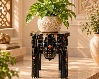 May include: A black wooden stool shaped like an elephant with hand-painted gold and colorful floral accents. The stool features a round top and four sturdy legs. A white ceramic planter with a gold leaf pattern holds a green pothos plant.
