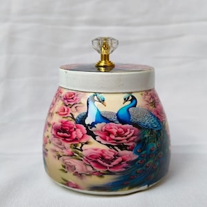 May include: A decorative jar with a lid, featuring a vibrant peacock and floral design. The jar's body is adorned with pink roses and two blue peacocks. The lid has a clear, crystal-like knob with a gold base.
