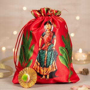 Puede incluir: Bolsa roja de satén con cordón, con una ilustración impresa de una mujer con vestimenta tradicional india. La bolsa presenta detalles de hojas verdes y ribetes dorados y verdes. Una roseta dorada con un pompón verde está en primer plano.