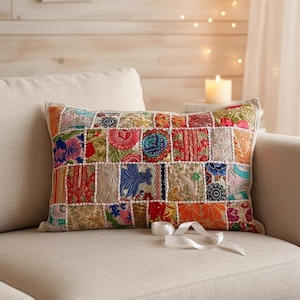 May include: A rectangular patchwork cushion with a variety of colourful fabric squares. The cushion features floral and geometric patterns in shades of red, blue, orange, and green. A white ribbon is placed on the sofa next to the cushion.