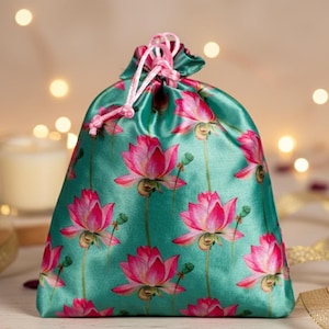 Op de afbeelding: Een turquoise satijnen trekkoordtas versierd met een herhaald patroon van roze lotusbloemen. De tas is vastgebonden met een roze trekkoord. De achtergrond is wazig met bokeh-lichten en een kaars.