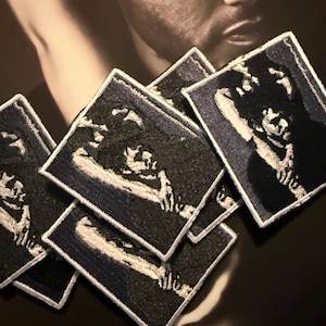 May include: Several black and white embroidered patches featuring a portrait design. Each patch has a dark blue border and a white stitched outline. The patches are displayed on a dark surface, with a blurred image of a person in the background.