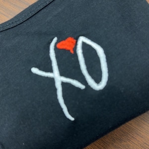 May include: Black face covering with white embroidered "XO" text and a red heart-shaped design. The face covering is made of a soft, dark fabric.