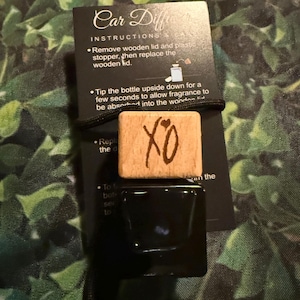 Puede incluir: Un ambientador de coche de madera con las letras "XO" grabadas en él. El ambientador está unido a una botella negra con una tapa negra. Las instrucciones para usar el ambientador están impresas en una tarjeta negra.