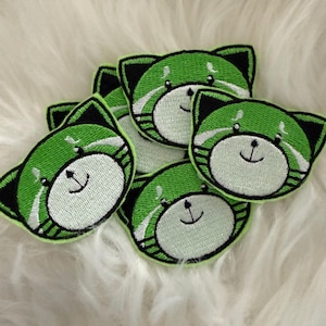 Könnte beinhalten: Fünf grüne gestickte Bügelbilder mit einem Comic-Tier mit schwarzen Augen und einem weißen Gesicht. Das Tier hat eine schwarze Nase und einen kleinen Mund mit einem Lächeln.