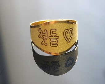 Puede incluir: Un anillo de oro con las palabras coreanas "처음" y un símbolo de corazón grabados.