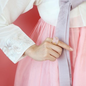 May include: A pink and white traditional Korean dress with a pink gemstone ring on the hand of the person wearing the dress.