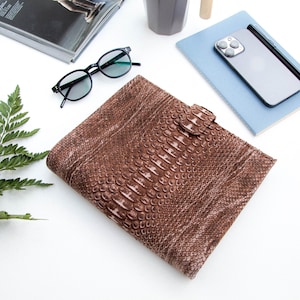 Peut inclure: Un carnet en cuir marron avec un motif en peau de serpent. Le carnet a une fermeture en métal et est posé sur une surface blanche.