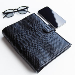 Peut inclure: Un portefeuille en cuir noir avec une texture de peau de reptile, un smartphone noir et des lunettes à monture ronde noires sont disposés sur une surface blanche. Le portefeuille a une fermeture à rabat. Accessoires.