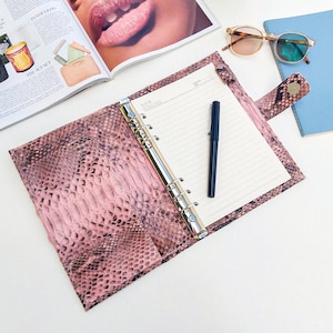 Peut inclure: Un classeur en cuir rose à motif peau de serpent avec un mécanisme de reliure à anneaux argentés et un stylo noir. Le classeur est ouvert pour révéler du papier blanc ligné.