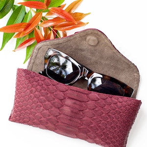 Puede incluir: Un estuche para gafas de sol de piel de serpiente burdeos con forro de ante marrón y un par de gafas de sol marrones en el interior.