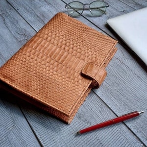 Peut inclure: Un carnet en cuir beige avec un motif peau de reptile, fermé par une patte assortie. Un stylo rouge, des lunettes et un ordinateur portable sont également sur la surface en bois.