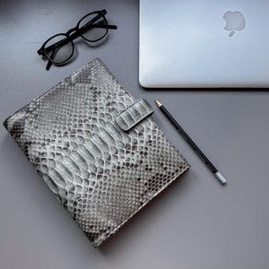 Peut inclure: Un ordinateur portable argenté, un carnet gris texturé imitation peau de serpent avec une fermeture assortie, des lunettes noires et un crayon noir sont disposés sur une surface grise. Le carnet est fermé.