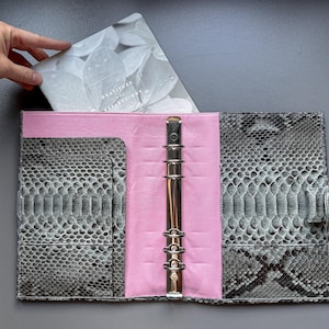 Peut inclure: Un agenda ouvert avec un extérieur à motif peau de serpent gris et un intérieur rose. L'agenda a un mécanisme de reliure argenté et une fermeture à bouton-pression. Une main place un livret avec le texte "Gratitude & Reflections" à l'intérieur.