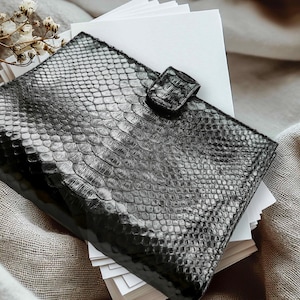 May include: A black leather wallet with a reptile skin texture, secured with a matching clasp. The wallet is resting on a stack of white paper and a beige linen surface. A sprig of dried flowers is in the background.