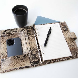 Peut inclure: Un agenda relié en cuir marron avec un motif peau de serpent. L'agenda est ouvert pour révéler une page lignée vierge et un stylo noir. Un smartphone noir se trouve dans la poche sur le côté gauche de l'agenda.