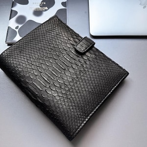 May include: Black leather notebook cover with a reptile skin texture. The cover has a flap closure and is lying on a light gray surface next to a notebook with a black and white pattern and the word "BELIEVE".