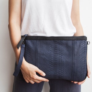 May include: A navy blue clutch bag with a snakeskin effect. The bag has a black zip and a wrist strap. The clutch is held by a person wearing a white t-shirt.