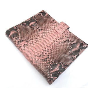 Peut inclure: Une couverture de carnet ou de tablette à motif peau de serpent rose et noir avec une fermeture à languette. La couverture est faite d'un matériau texturé et a une forme rectangulaire. Le schéma de couleurs est un mélange de rose, de noir et de marron.