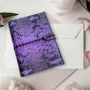 May include: A purple and black snakeskin-patterned journal with a black elastic closure. The journal is on a white envelope and a light pink envelope, with white flowers and petals in the background.