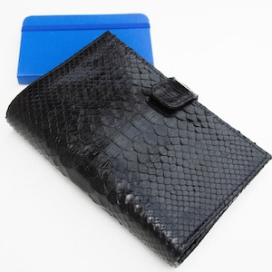 Peut inclure: Un portefeuille en cuir noir avec un motif en peau de serpent et un fermoir argenté. Le portefeuille est fermé et posé sur une surface blanche.