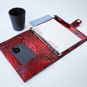 Peut inclure: Un agenda en cuir rouge à motif peau de serpent, ouvert pour révéler un carnet ligné vierge, un stylo noir et un smartphone noir.