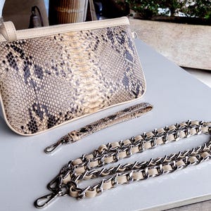 Può includere: Una pochette beige in pelle di serpente con una tracolla a catena argentata. La borsa ha una chiusura a lembo e una piccola tasca sul davanti.