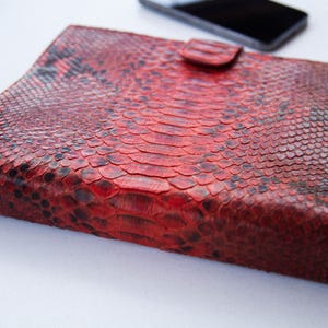 Peut inclure: Pochette en peau de serpent rouge et noir avec fermeture à rabat. La pochette en cuir présente un motif texturé et écailleux et une petite fermeture à languette. Un smartphone noir est visible en arrière-plan.