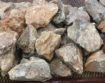 May include: A pile of various-sized rocks in shades of brown, gray, and tan. The rocks have rough, textured surfaces and are resting on a metal mesh surface. The rocks vary in size and shape, creating a natural, earthy aesthetic.