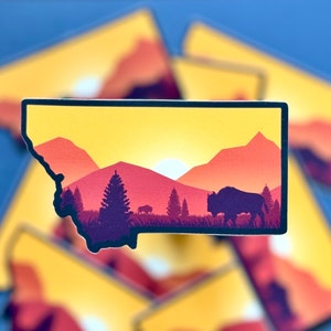 May include: A silhouette of a bison standing in a field with a sunset in the background. The silhouette is in front of a mountain range with trees in the foreground. The image is a sticker with a black outline.
